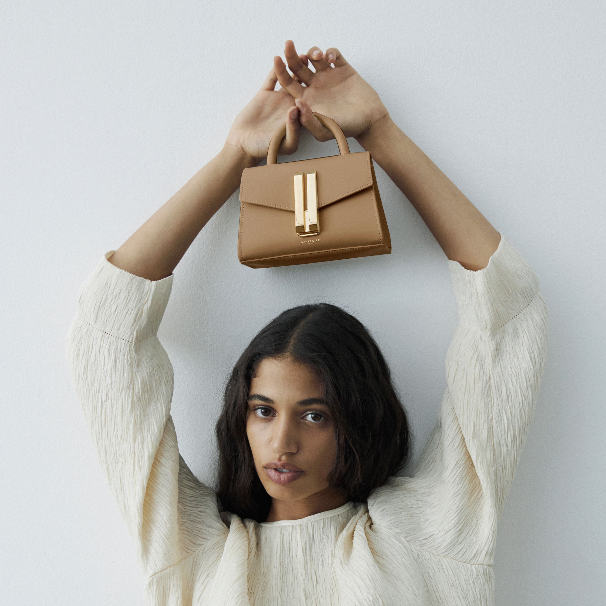 Model posing, holding the toffee Demellier handbag above her head. Model is wearing a white top.