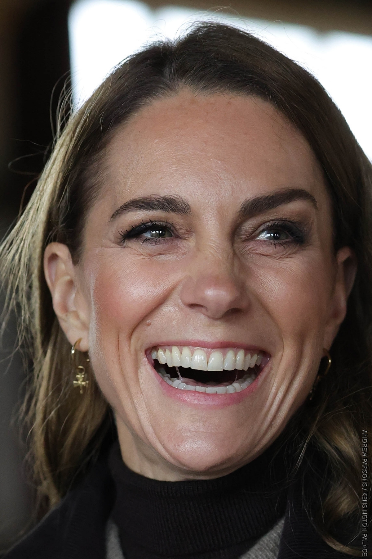 A super tight shot of Kate Middleton's face, she is laughing and looking to the side.
In her ears, are the Daniella Draper gold hoop earrings and Shamrock charms.