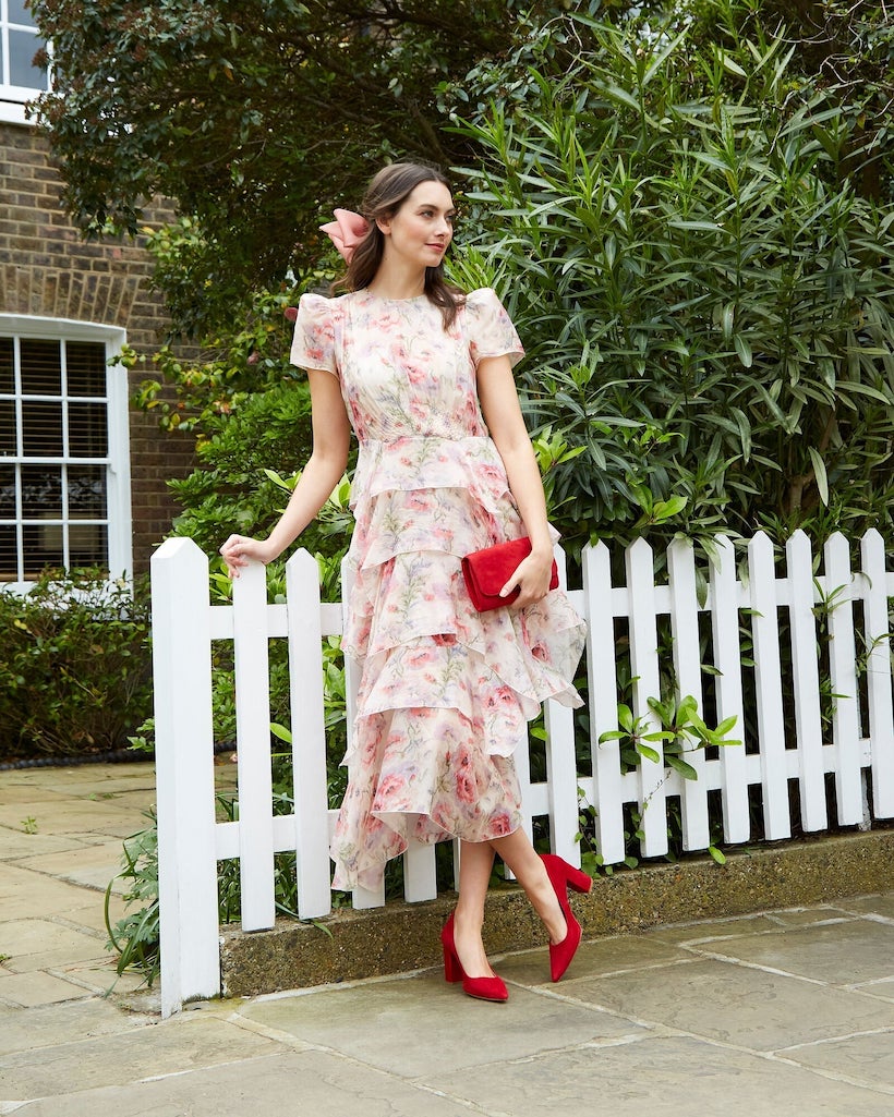 A woman wearing a flowing floral dress with pink and red tones stands beside a white picket fence, holding the Emmy London Natasha clutch in red suede. She pairs the bag with matching red heels for a soft, romantic look.