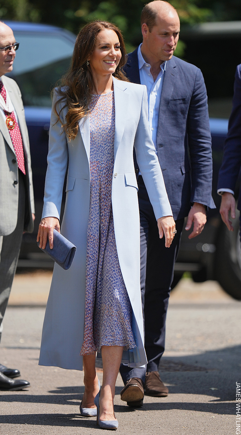 Kate Middleton walking outdoors, wearing the Emmy London Rebecca Court Shoes in Riviera Blue Suede, paired with a light blue coat, lilac printed dress, and a matching clutch bag.