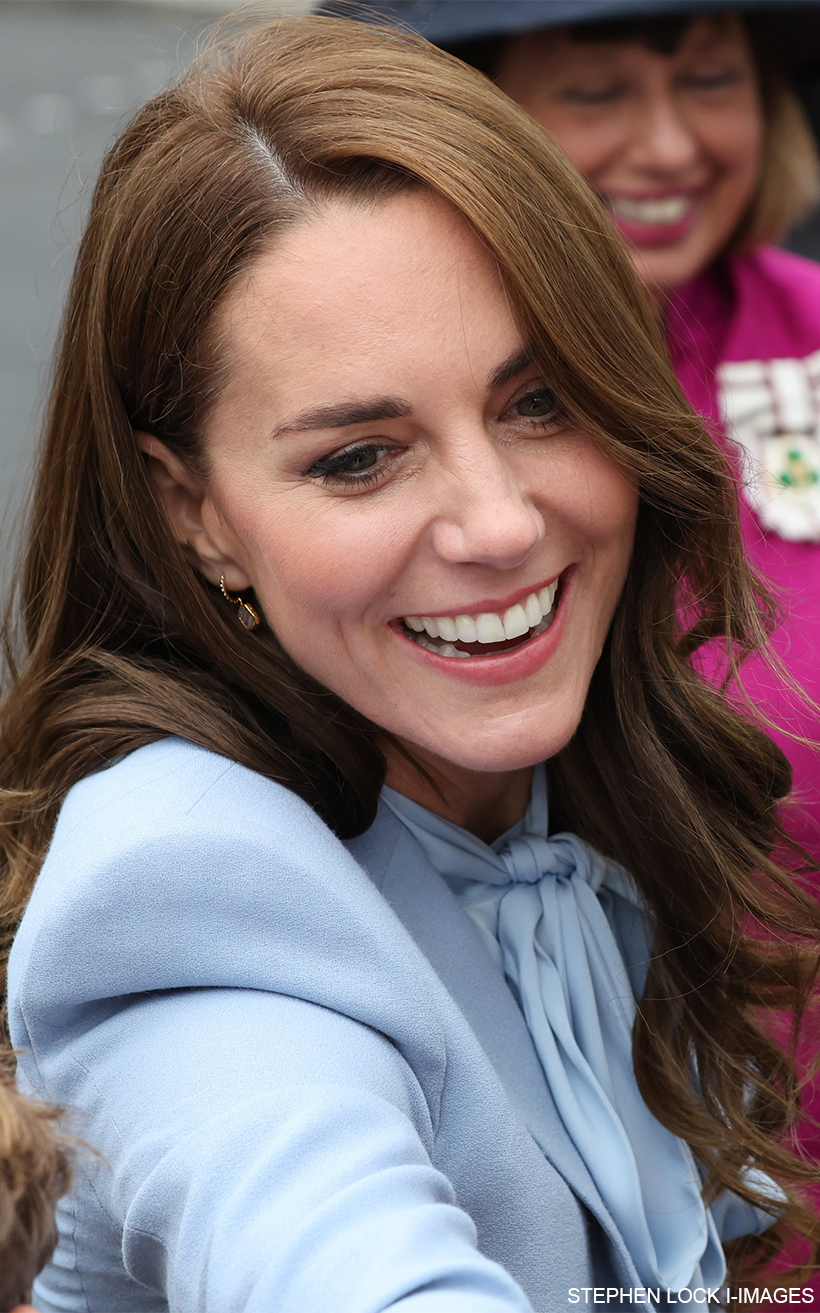 Close up of Kate Middleton smiling and greeting a member of the public in the crowd. She's wearing her blue coat, blue bow blouse and pink/gold earrings.