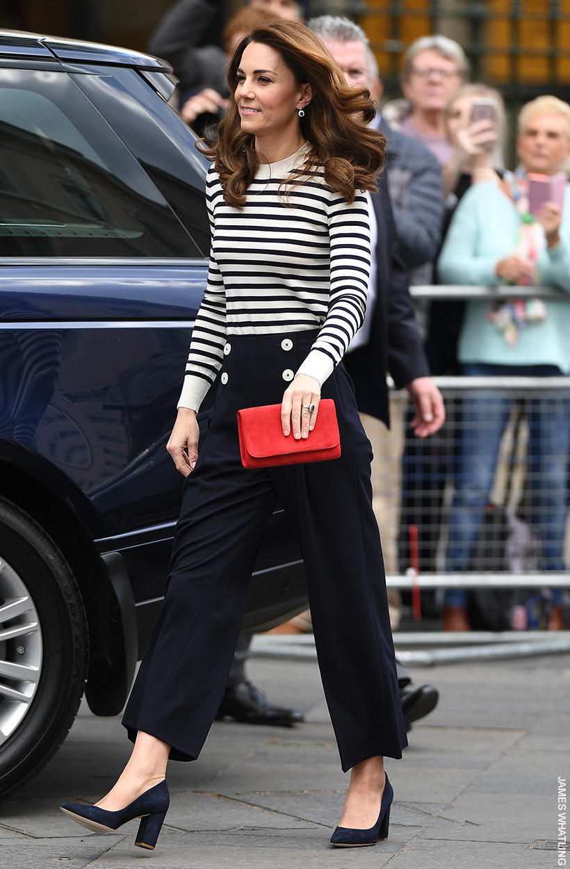 The Princess of Wales walking, wearing a striped jumper and black trousers. She is holding a vivid red clutch bag in her hand.  It's from the British brand Emmy London. 