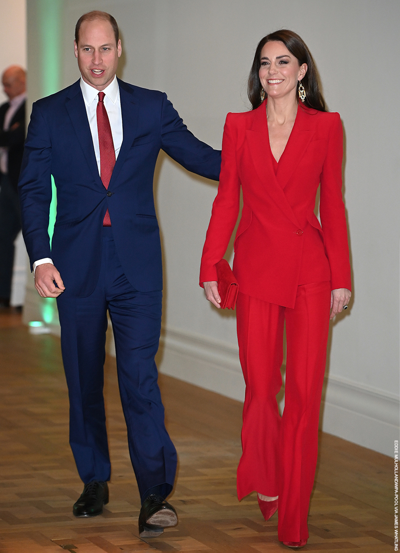 Kate Middleton, smiling and walking with Prince William. The Princess is wearing a bright red suit by Alexander McQueen.