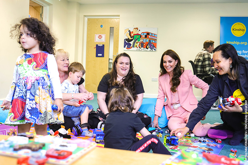 Kate Middleton playing with children at a Kinship support group. She's wearing a pink suit with pearl accessories.