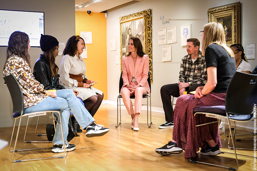 Kate Middleton sitting down with a group of people inside the Foundling Museum. She's wearing a pink suit with pearl accessories.