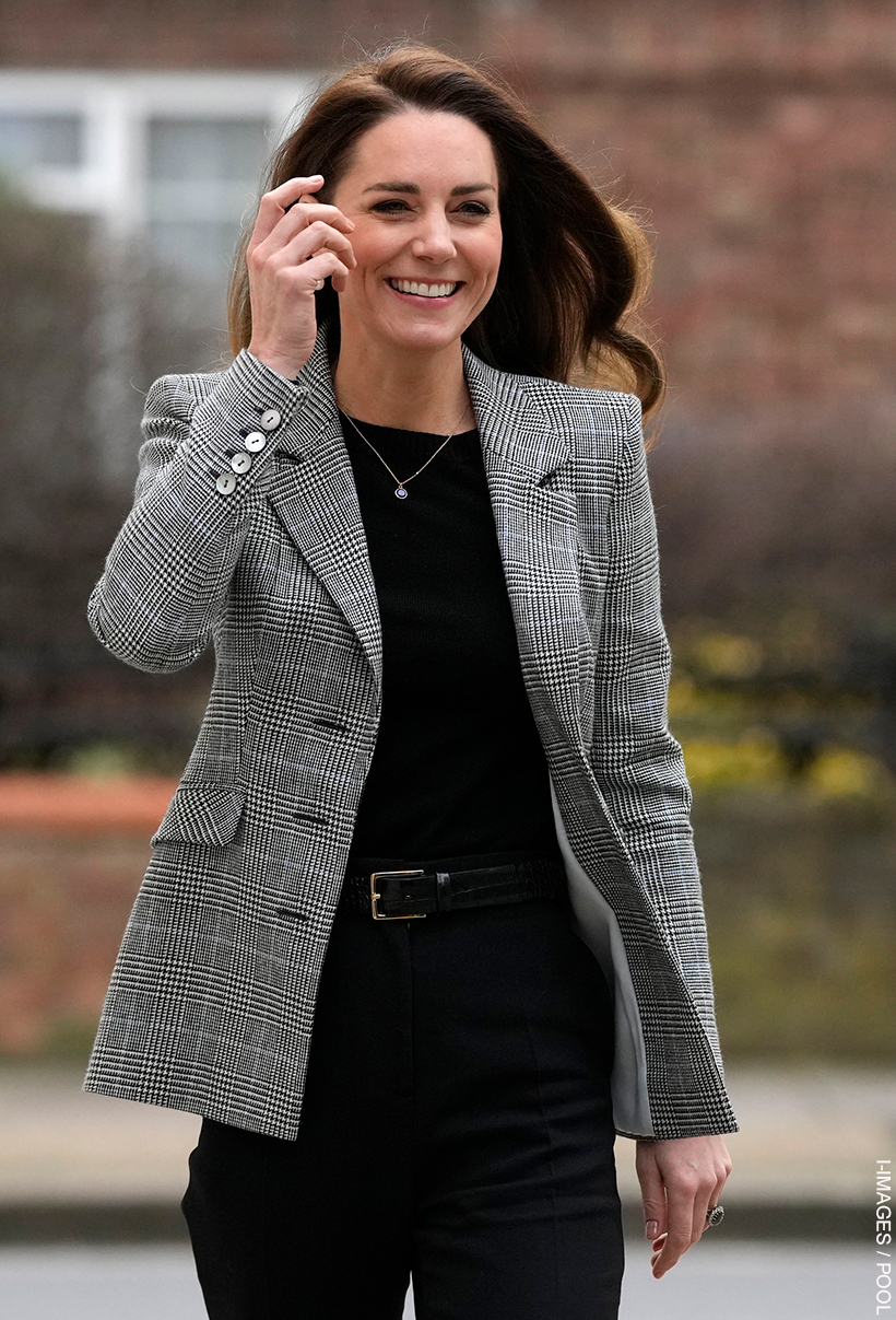 Kate Middleton wearing the glen check blazer over a black sweater. She is smiling and her hair is blowing in the wind.