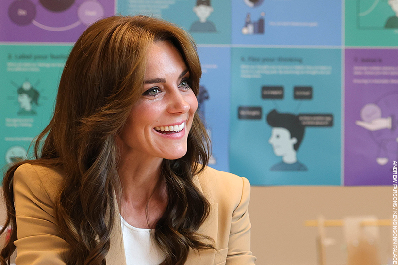Kate Middleton smiling and engaging in conversation with somebody off camera. The colourful blue and purple background contrasts with her camel suit and dark curly hair.