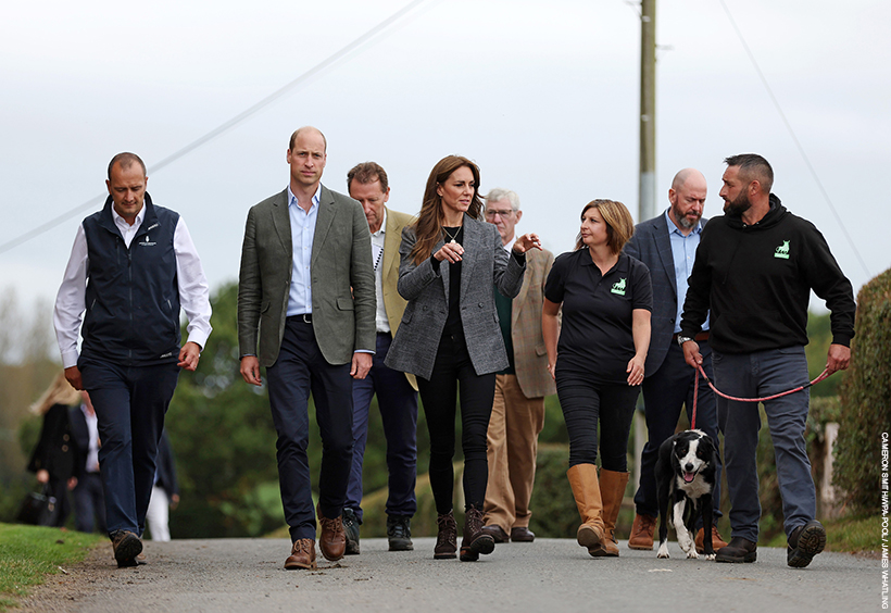 William and Kate stand in the middle of a crowd of people, walking and talking. Kate is gesturing as she talks. One of the charity owners, a farmer, walks a black and white collie dog.