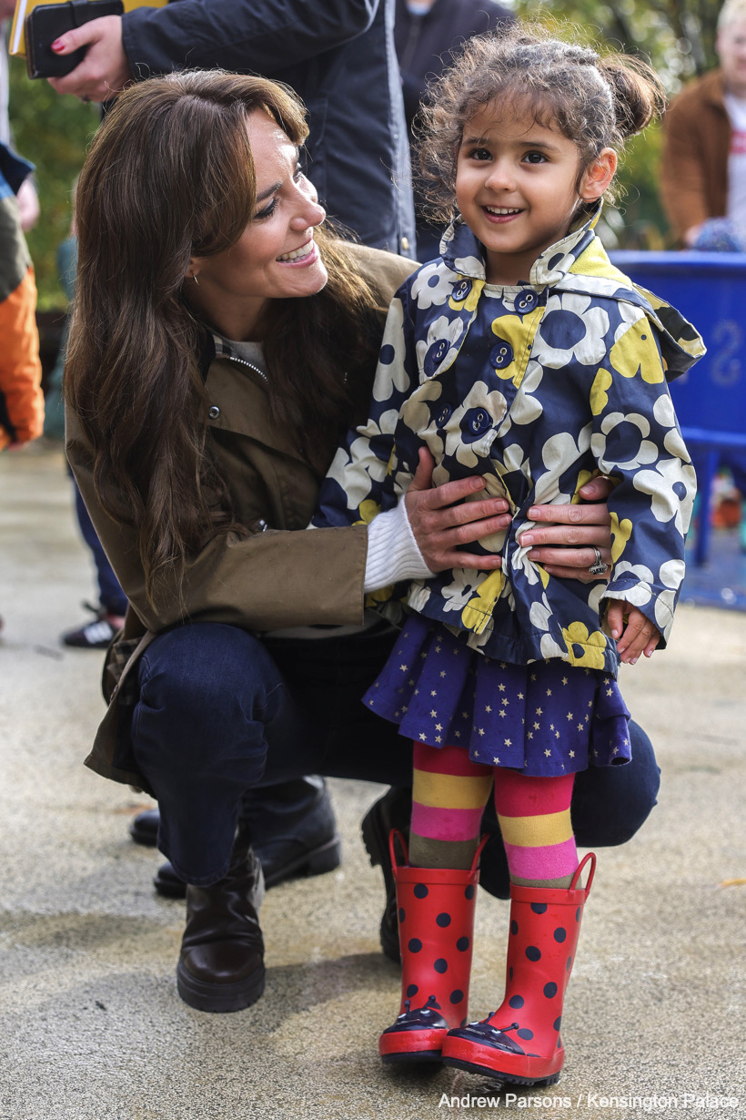 Kate crouches on the floor and cuddles a little girl dressed in a flowery raincoat, star print skirt, rainbow tights and ladybird wellies.