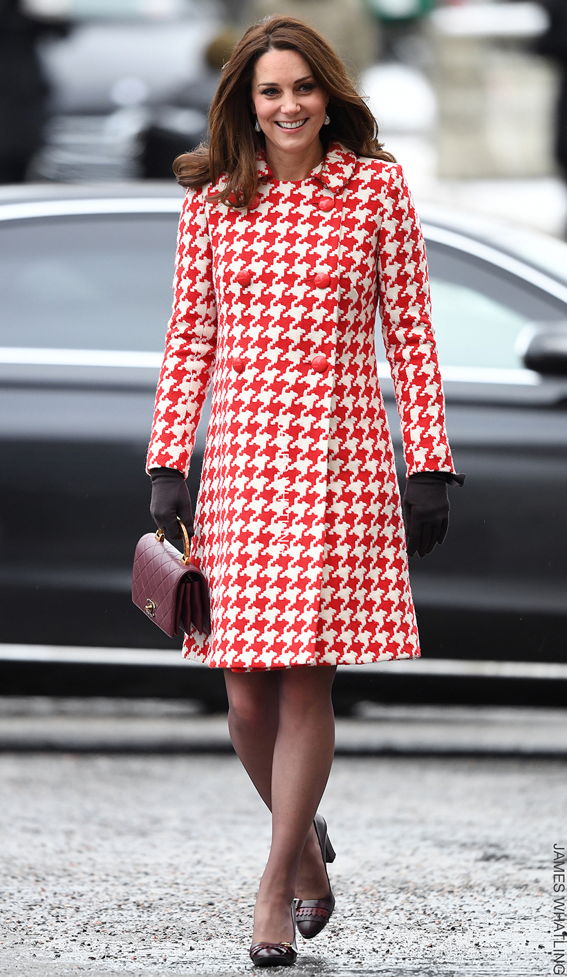 Kate Middleton, 6-7 months pregnant with Prince Louis during her visit to Stockholm, Sweden in 2018, wearing a red and white houndstooth coat by Catherine Walker & Co, paired with black gloves, a maroon handbag, and maroon heels.