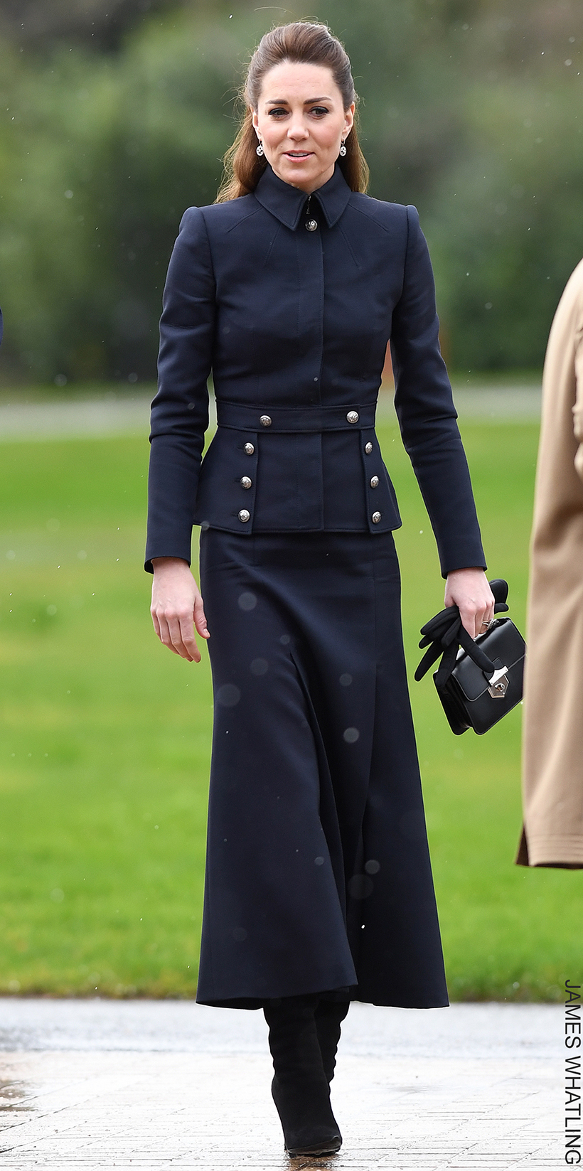 Kate Middleton dressed in a navy military-style jacket with silver buttons, paired with a matching midi skirt, black heeled boots, and carrying a black handbag, walking outdoors.