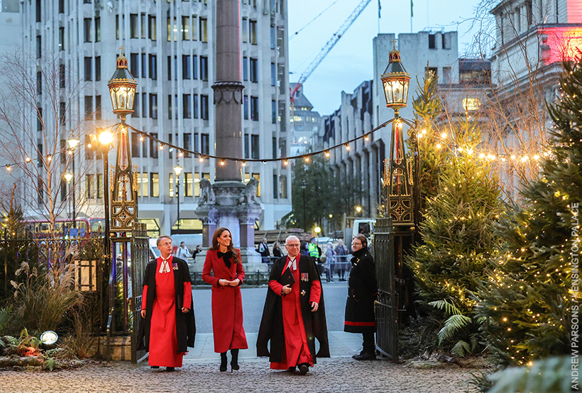 Kate Middleton wears a long red coat with a black velvet bow as she waves and walks into the 'Together At Christmas' Carol Service being held at Westminster Abbey. She walks with two clergymen.
