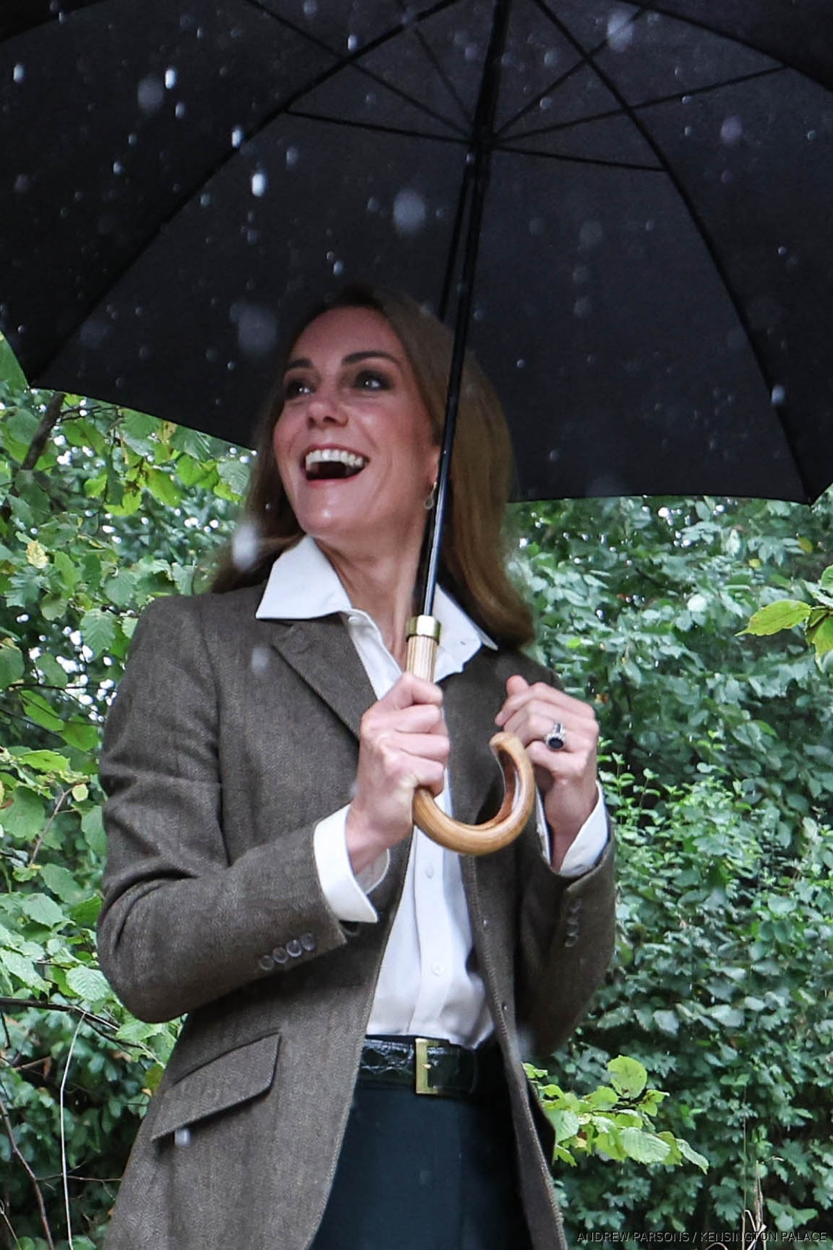 The Princess of Wales smiles looking up while holding an umbrella, she is wearing a tweed jacket and underneath is a crisp white shirt.