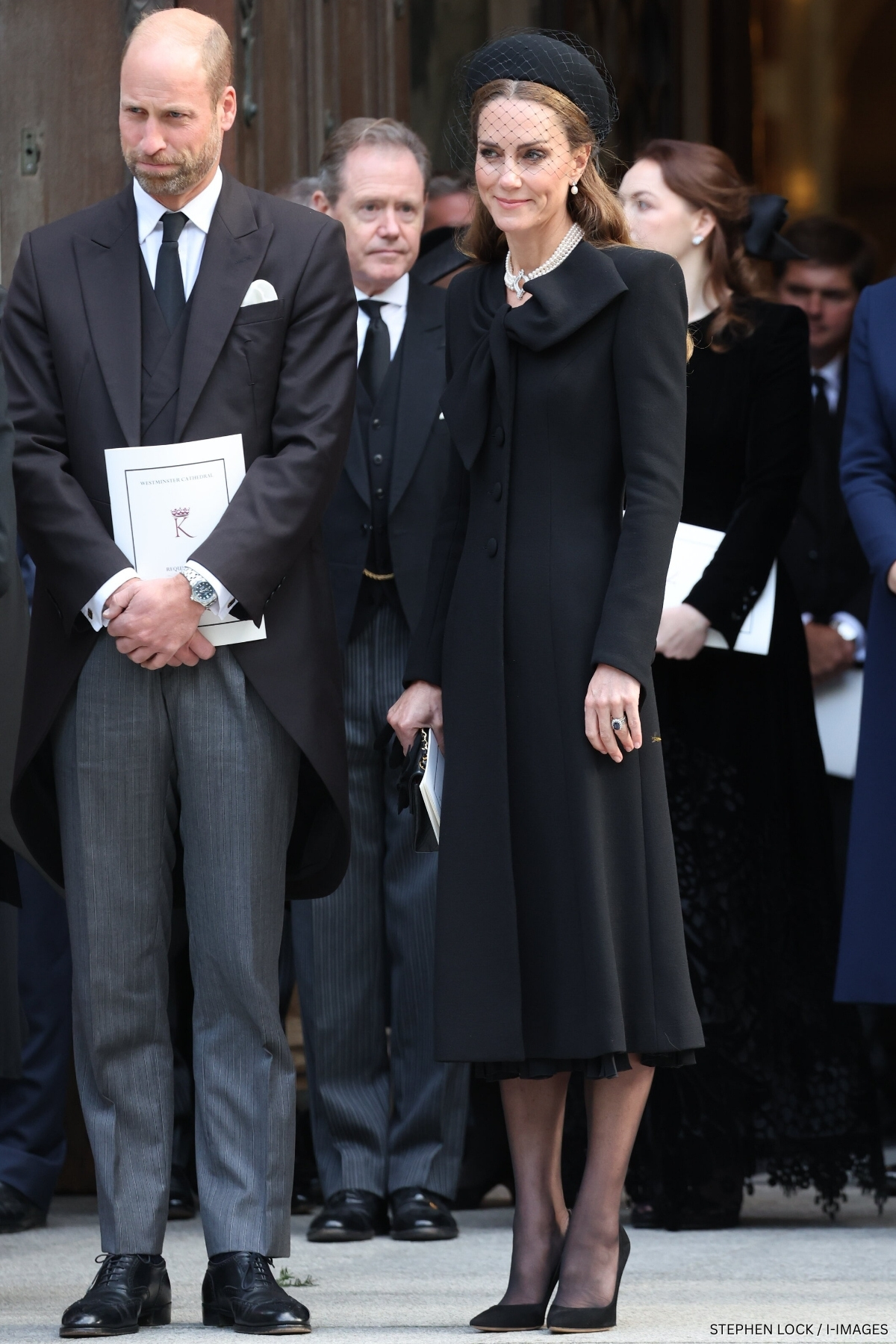 The Princess of Wales in a black coat dress with a beau tie detail at the neckline, black pillbox hat with veil and black quilted chanel handbag.
