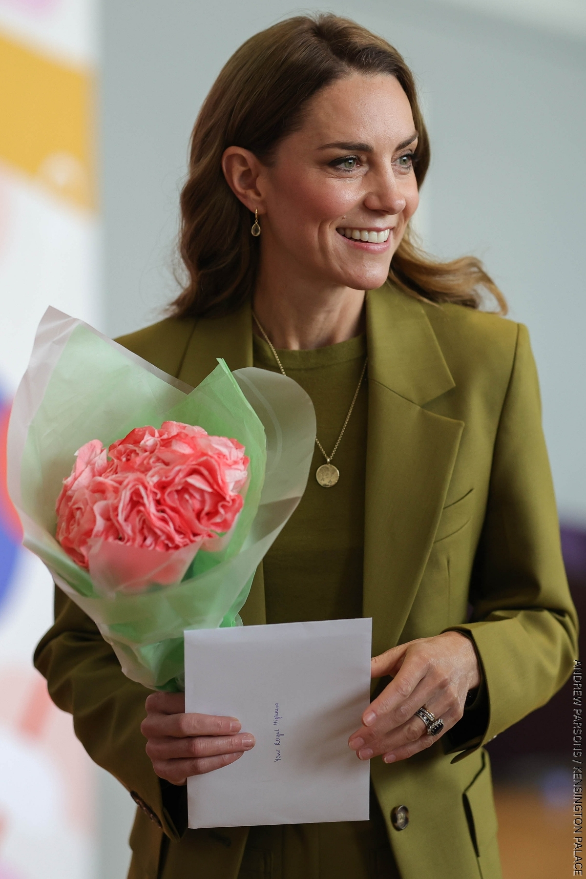The Princess of Wales holding a bunch of flowers, she is wearing an olive green suit and a gold pendant necklace by Daniella Draper.