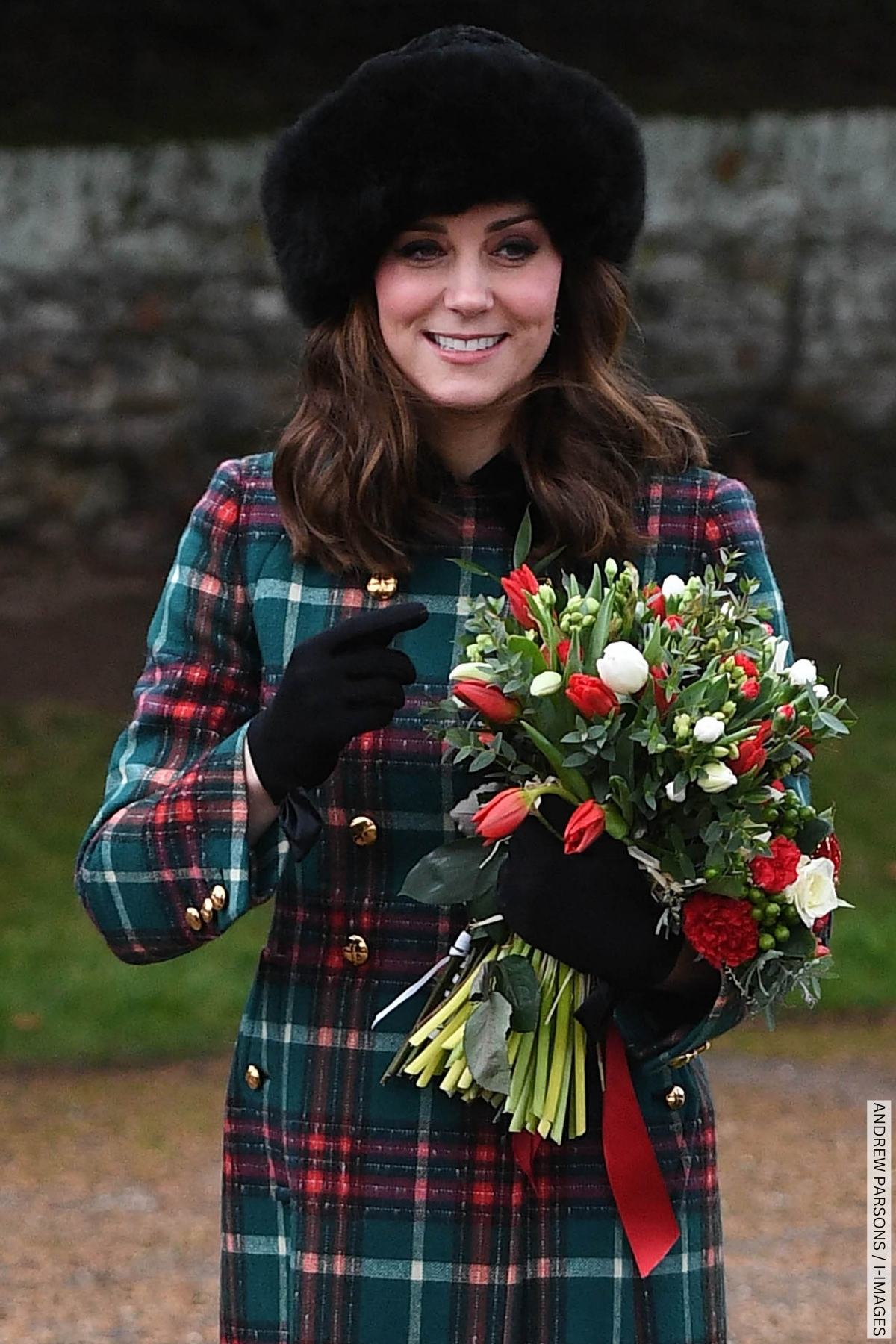 Kate Middleton wears a Christmassy green and red check coat, black fur hat and black gloves; she's holding a bunch of flowers