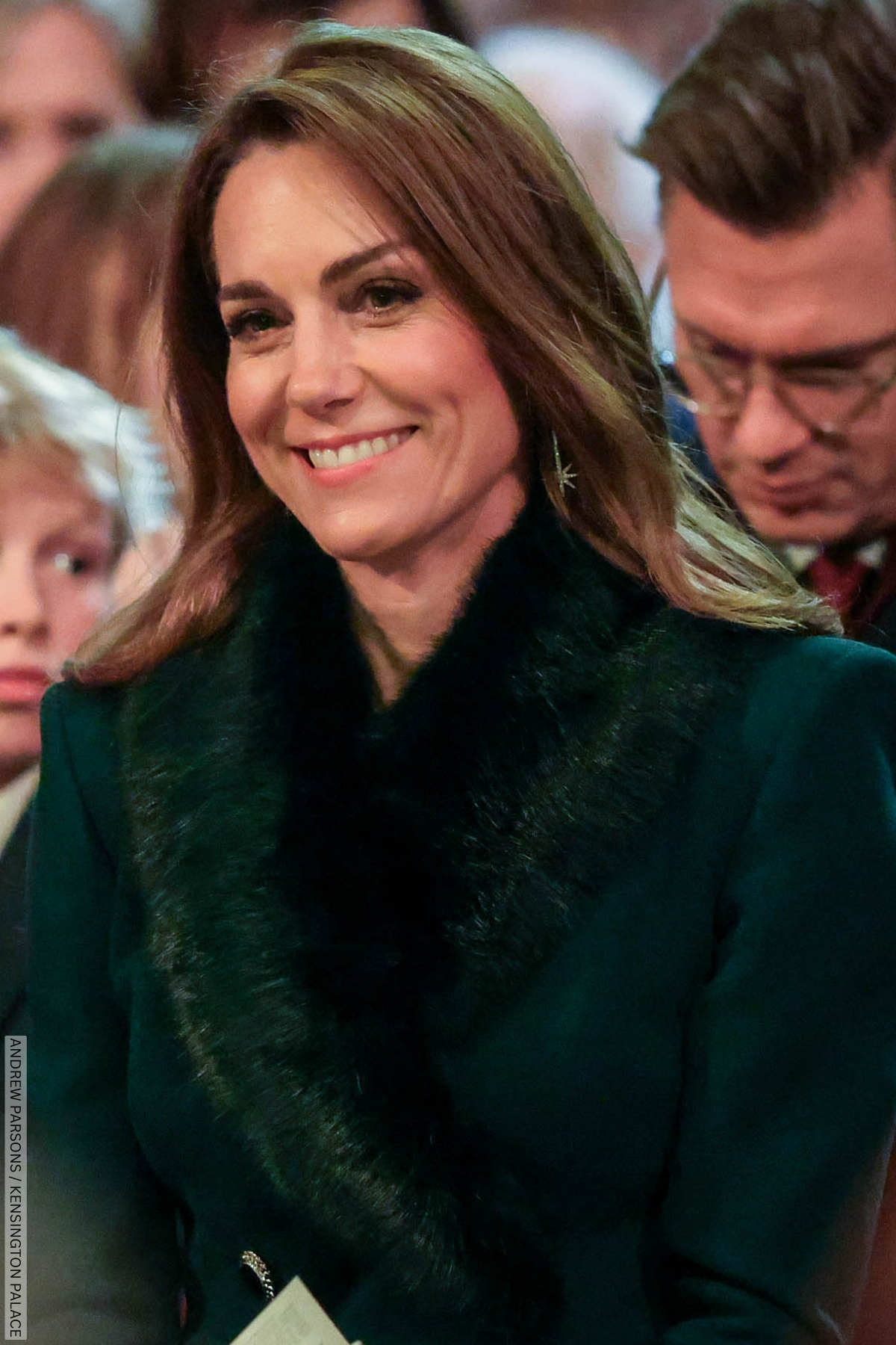 The Princess of Wales sat smiling during her carol concert. She wears a dark green fur lapel on the top of her coat