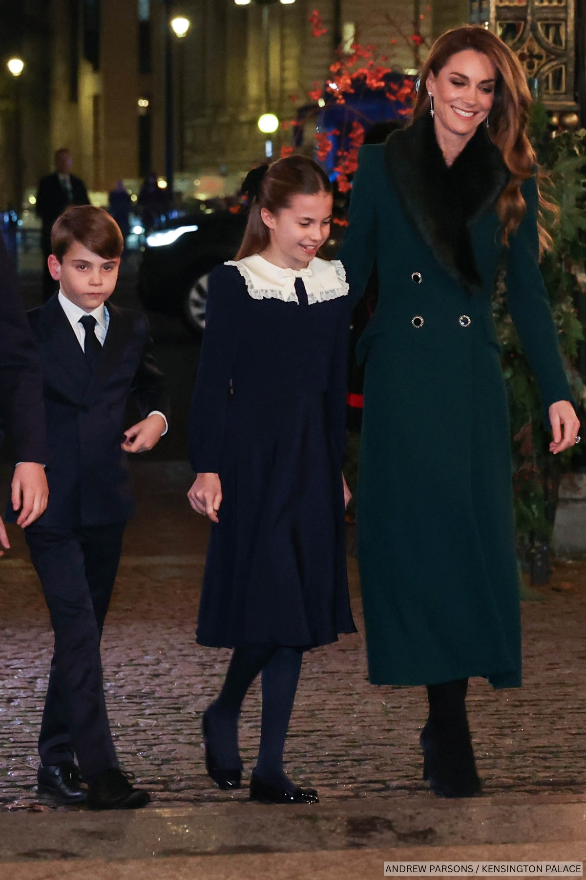 The Princess of Wales walking into the 2025 Together at Christmas Carol service, with two of her children. The Princess wears a deep green coat with fur lapel, black boots and a pair of sparkling star earrings.